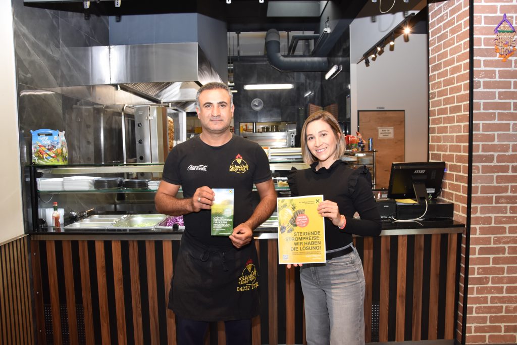Hier stehen zwei Personen in einem Kebaphaus und halten zwei Flyer in der Hand. 