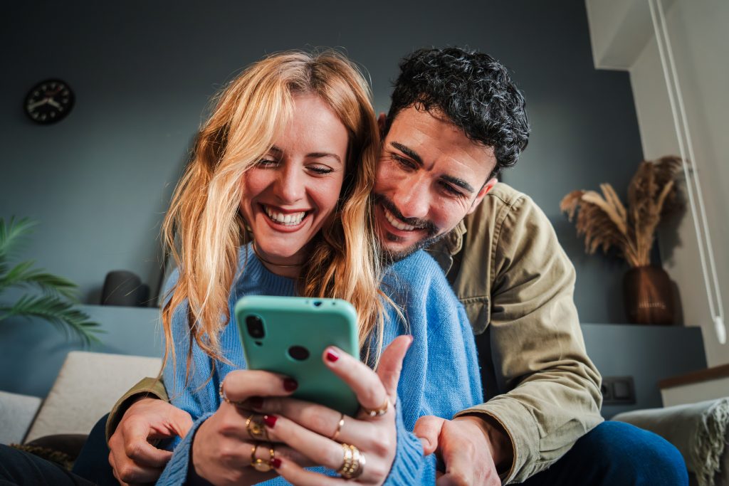 Young caucasian couple smiling using a cellphone to do shopping online. Man and woman watching the social media with a smartphone. Adult friends having fun browsing on internet with a mobile phone. High quality photo