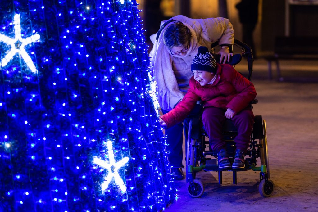 Happy child in a wheelchair touching a big illuminated christmas tree with his grandmother at night in a city