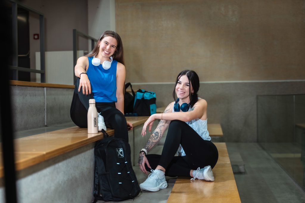 Zwei Mädels sitzen in einer Turnhalle in Sportbekleidung und lächeln in die Kamera