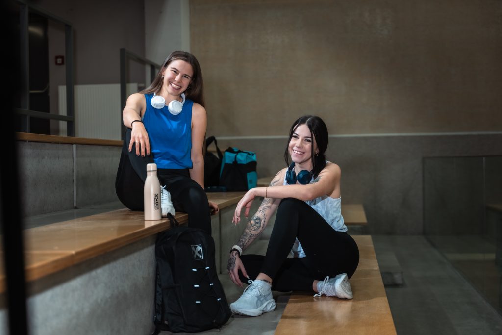 2 Mädels sitzen im Sportoutfit in einer Turnhalle und lächeln in die Kamera