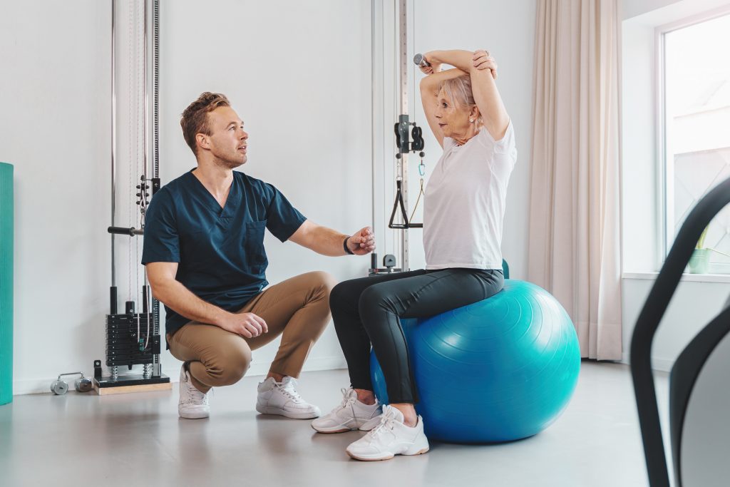 Senior woman sitting on a Pilates ball in gym while personal trainer instructs on exercise routine. Both smiling and engaged in the workout.