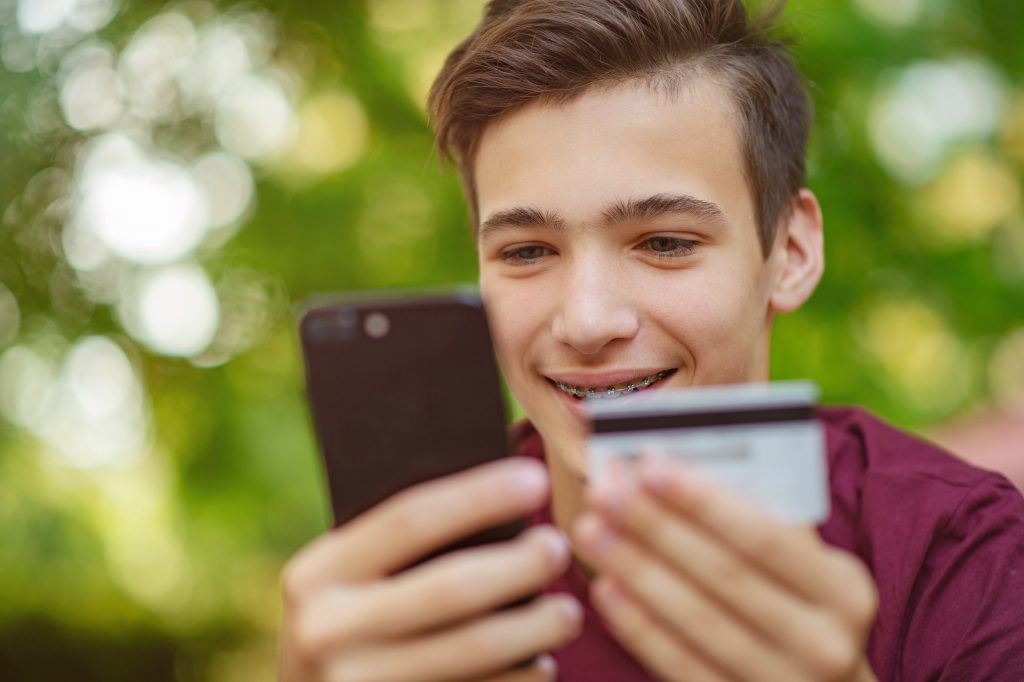 Teenage boy with a credit card and phone makes purchasing outdoors. Happy young man is using smart phone and bank card for online shopping. Handsome smiling white guy holds bank card and cell phone