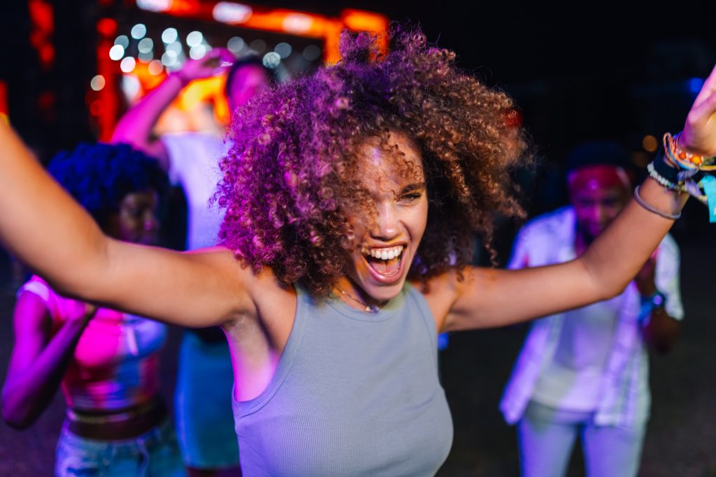 Young woman with curly hair enjoys the vibrant atmosphere of a music festival, dancing with energy and excitement alongside her friends under colorful neon lights