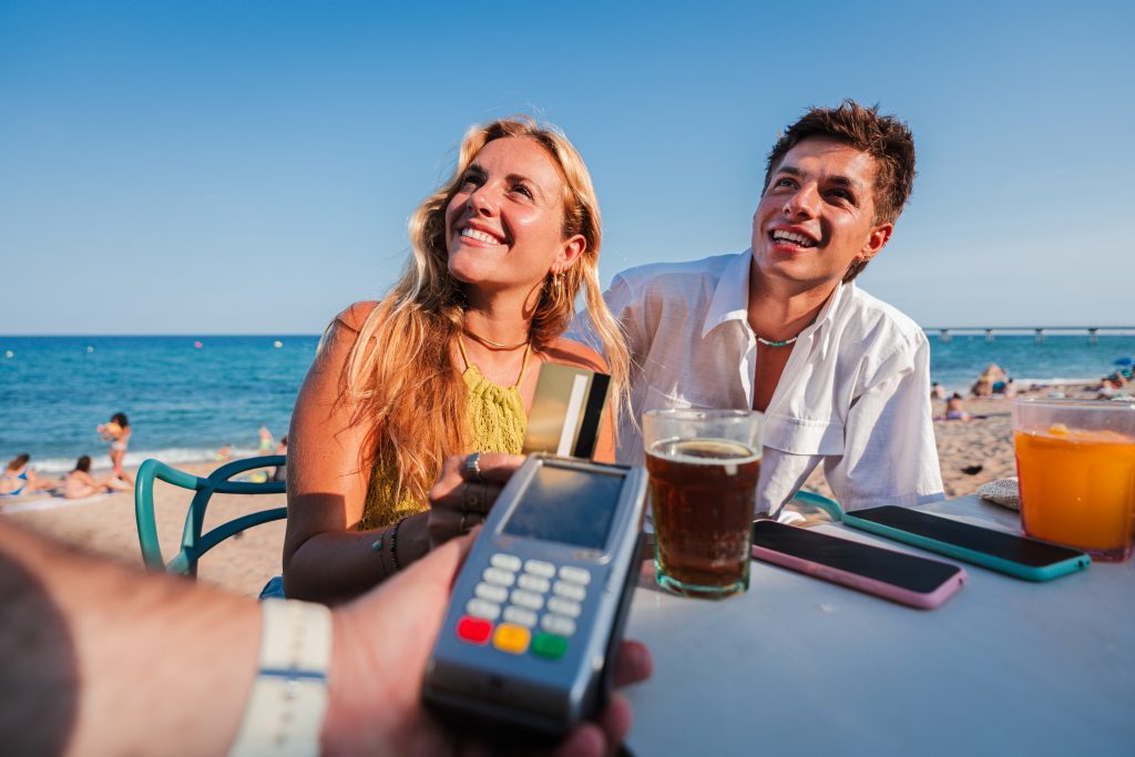 Young Caucasian couple sitting on the terrace of a beach bar, paying the bill with a credit card to the cashier.Two people giving a payment to the waiter after a good service. Client checkout at shore. High quality photo