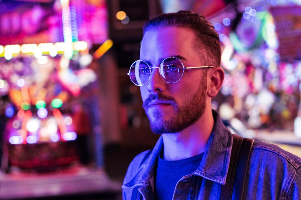 Ein junger Mann im Neon-Licht is hier zu sehen. Er trägt eine Brille. 