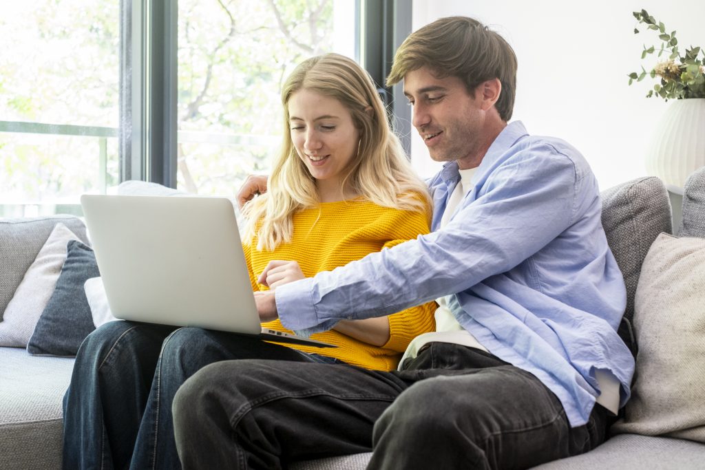 Young smiling couple relaxing on a comfortable sofa at home, sharing a laptop and browsing online, enjoying leisure time and modern connectivity